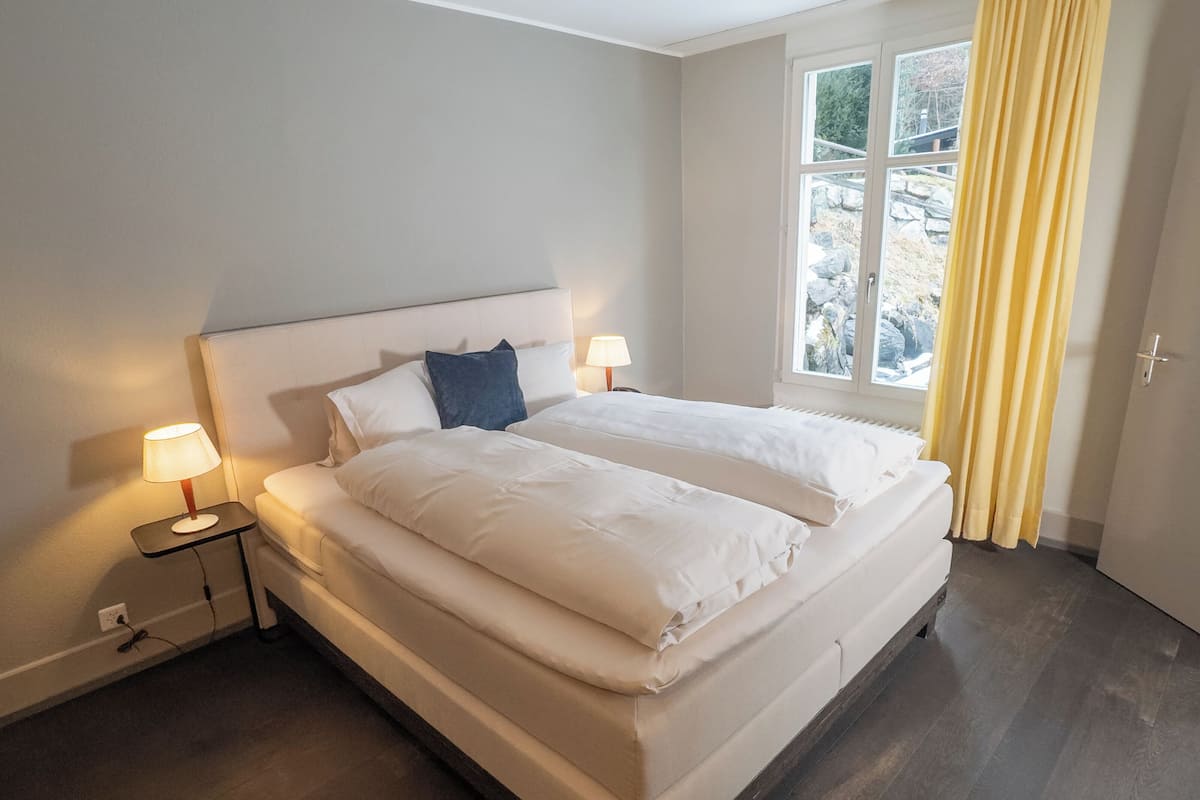 Cama doble con sábanas blancas, dos mesitas de noche con lámparas, una ventana con cortinas y vistas a rocas y árboles.