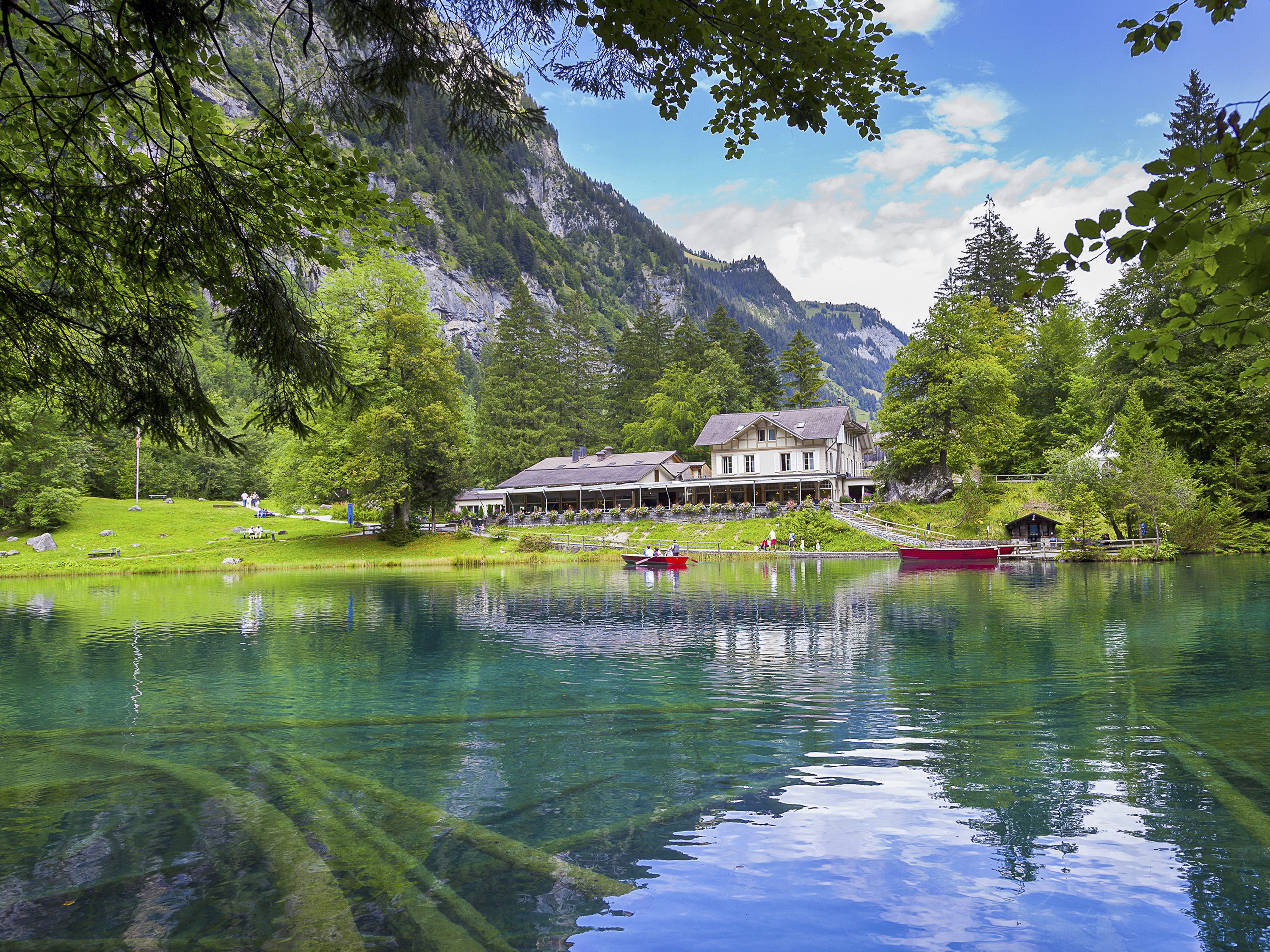 Un hôtel situé au bord d’un lac, avec un grand bâtiment, un quai avec des bateaux et une chaîne de montagnes en arrière-plan.