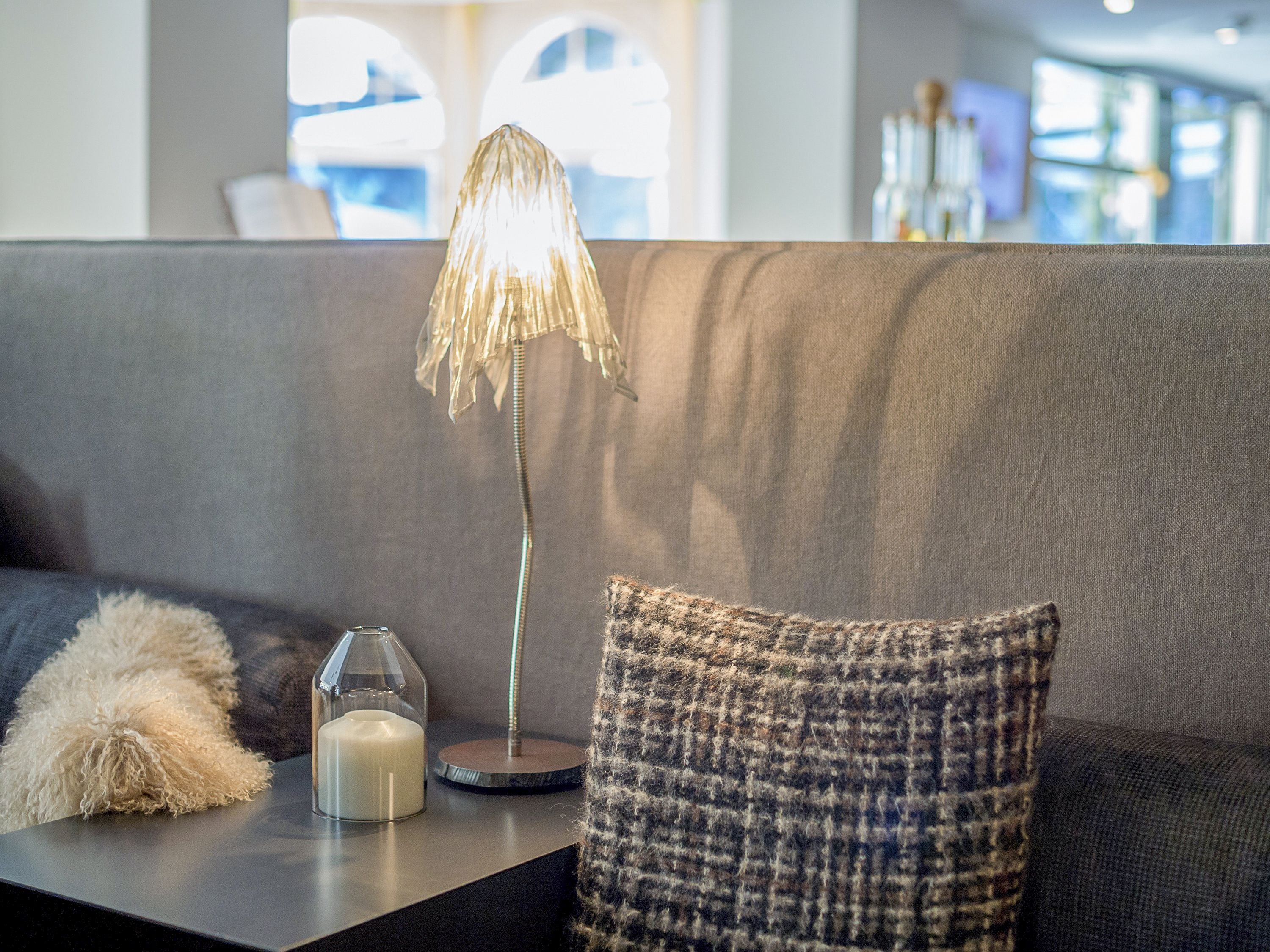 Ett modernt vardagsrum med en grå soffa, en mönstrad prydnadskudde, ett ljushållare i glas och en unik lampa med en metallisk fot.