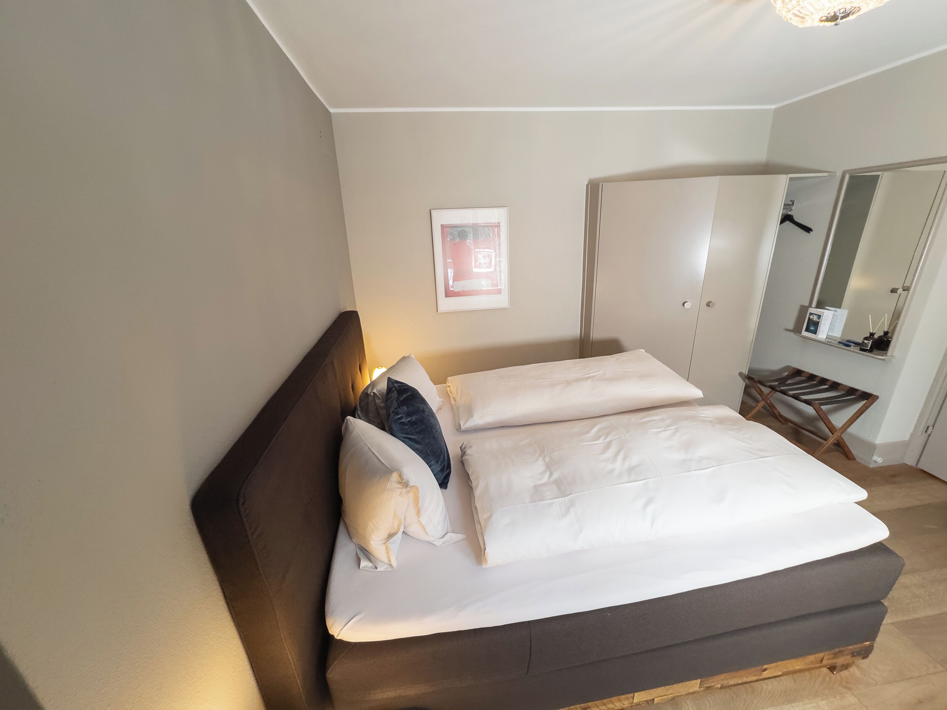 Un dormitorio con cama, almohadas, una mesita de noche, un espejo y un cuadro en la pared.