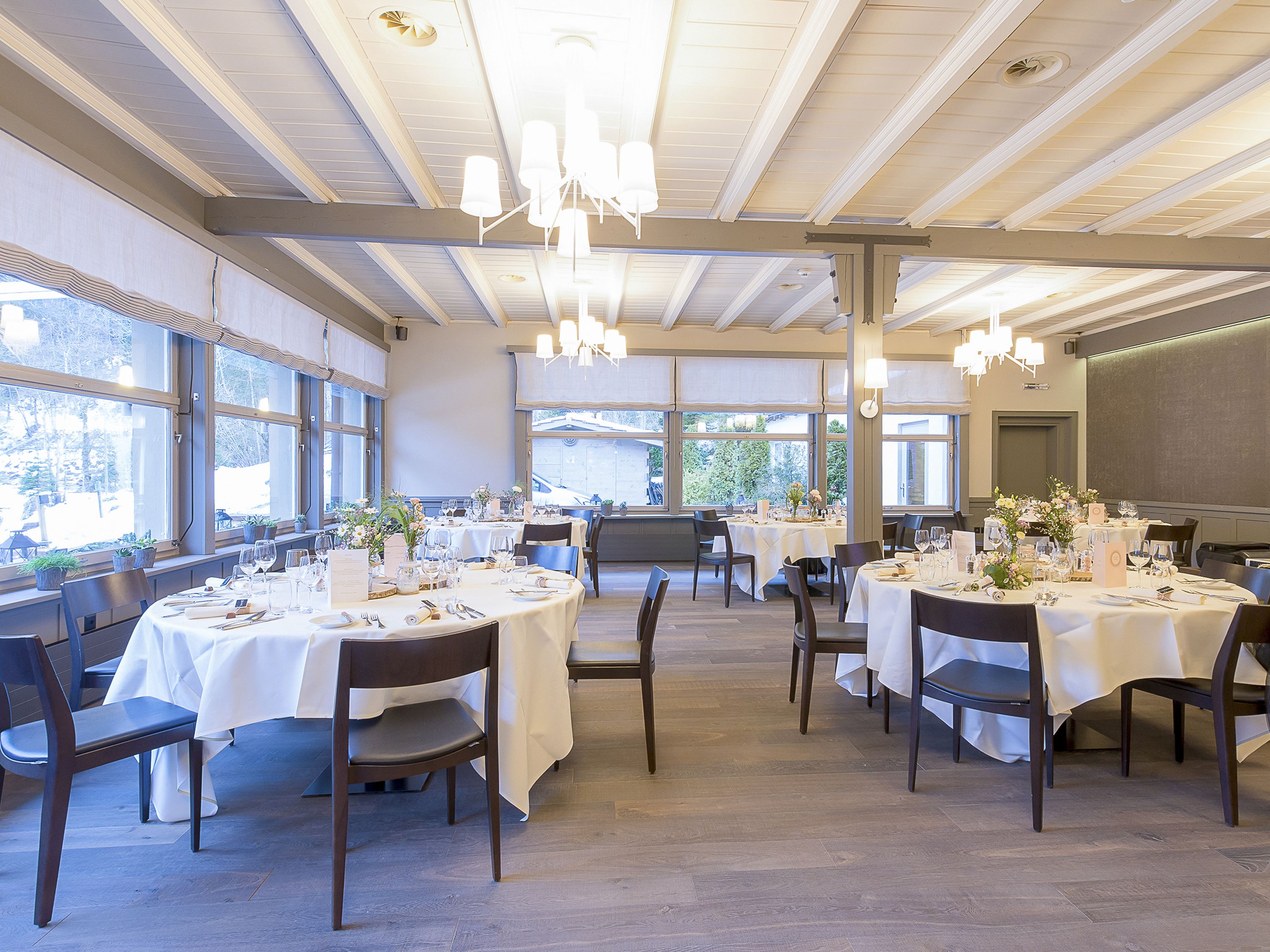 A dining area with multiple tables set for a meal, large windows, and a high ceiling with chandeliers.