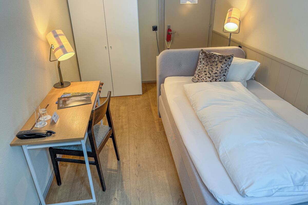 Habitación de hotel pequeña con una cama individual, un escritorio con silla, una lámpara y una puerta con un letrero.