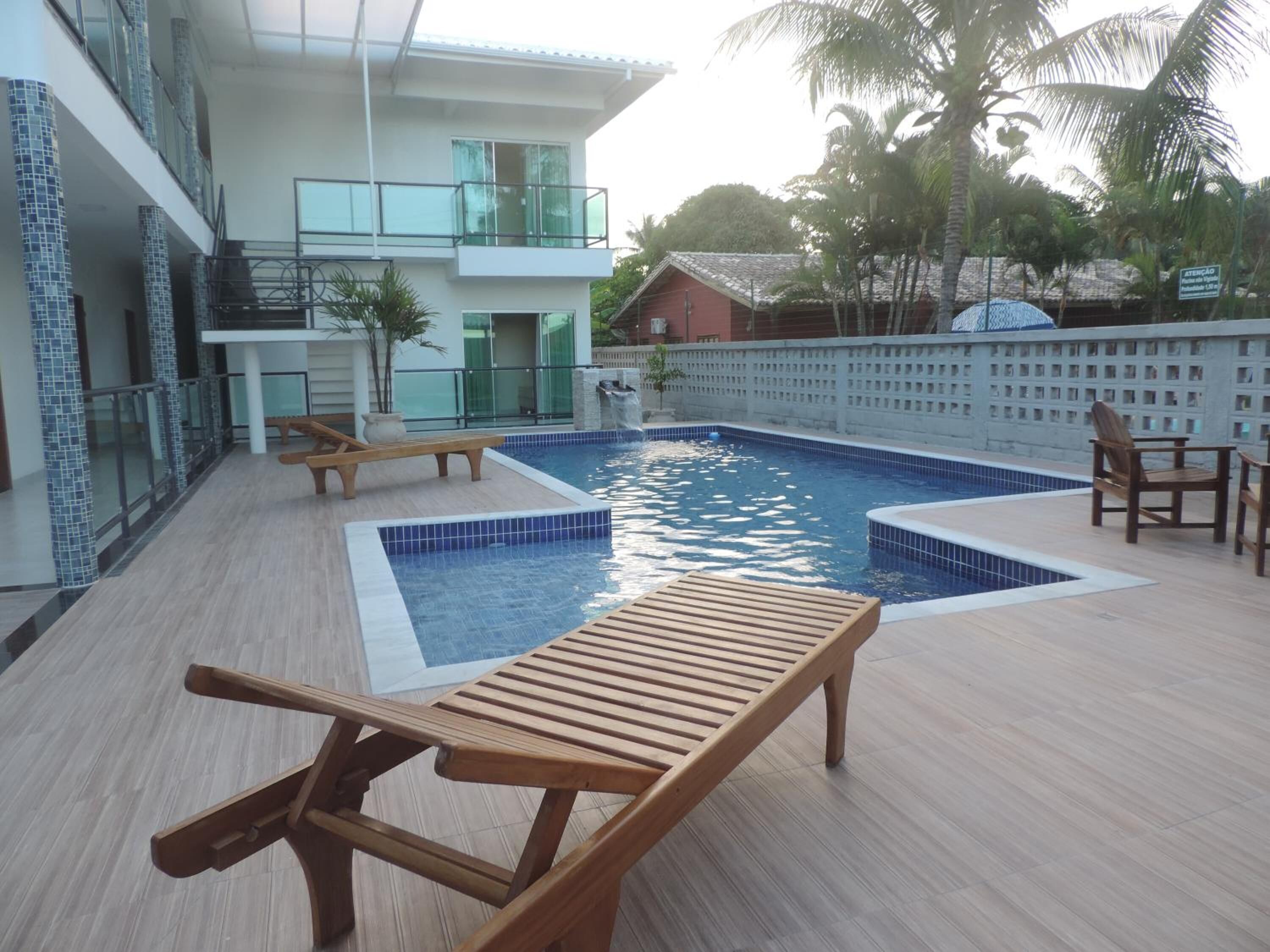 Una piscina con sillones de descanso, un edificio con balcón y palmeras al fondo.