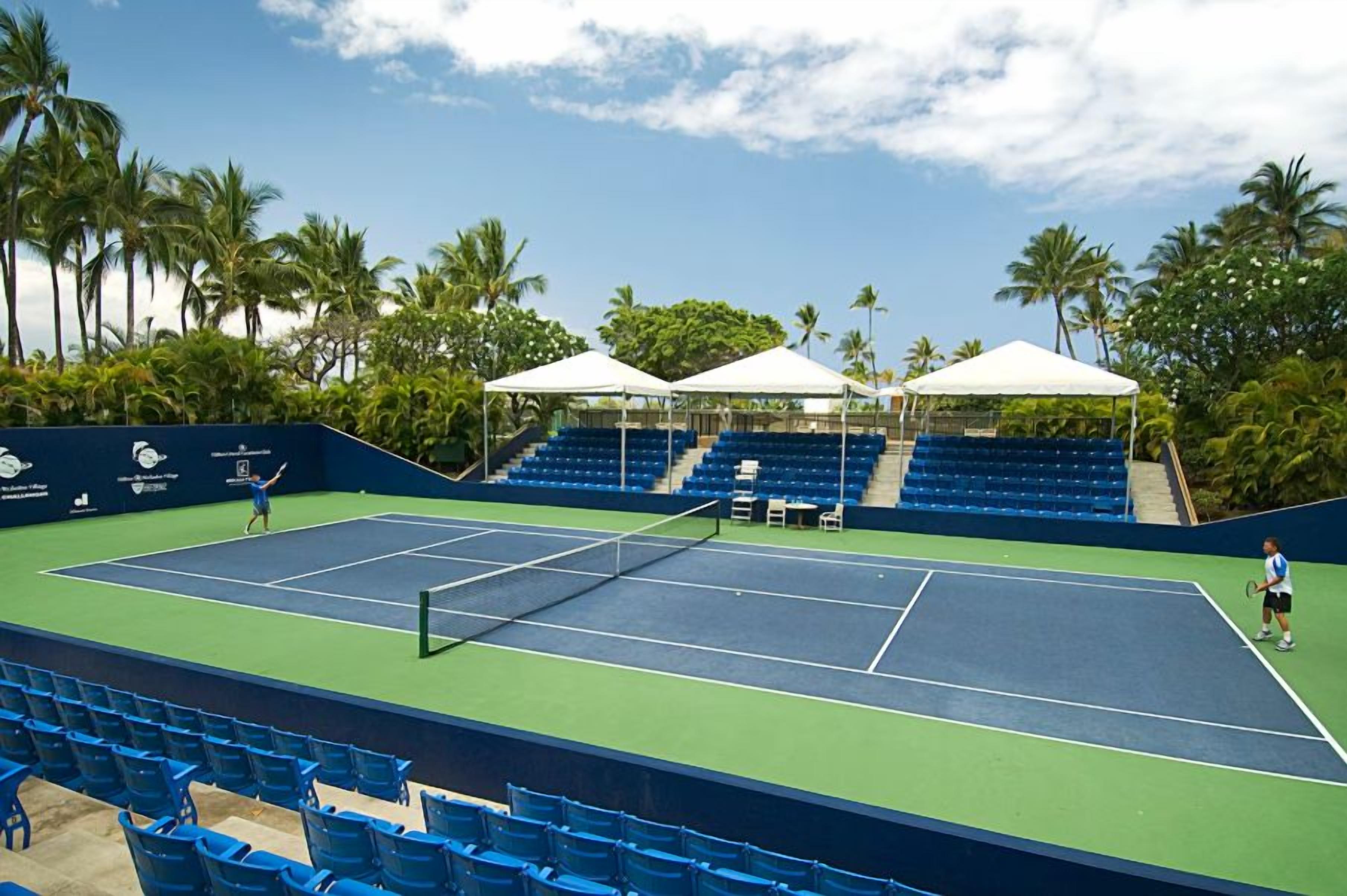 Cancha de tenis con asientos azules y toldos blancos, rodeada de palmeras.