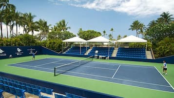 Cancha de tenis con asientos azules y toldos blancos, rodeada de palmeras.