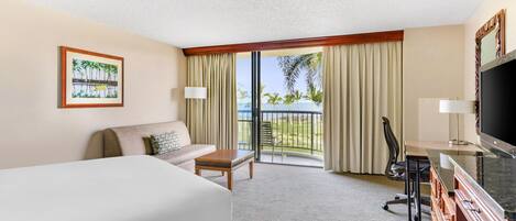A hotel room with a large bed, a desk with a flat-screen TV, a chair, a sofa, and a balcony with a view of palm trees and the ocean.
