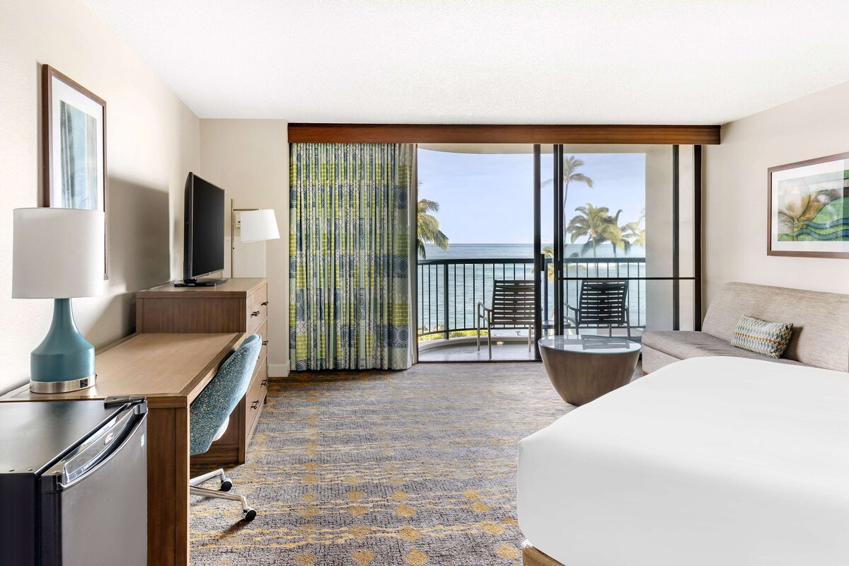 Une chambre d’hôtel avec un lit, un bureau doté d’une lampe, une télévision, un balcon offrant une vue sur l’océan et une moquette à motifs.