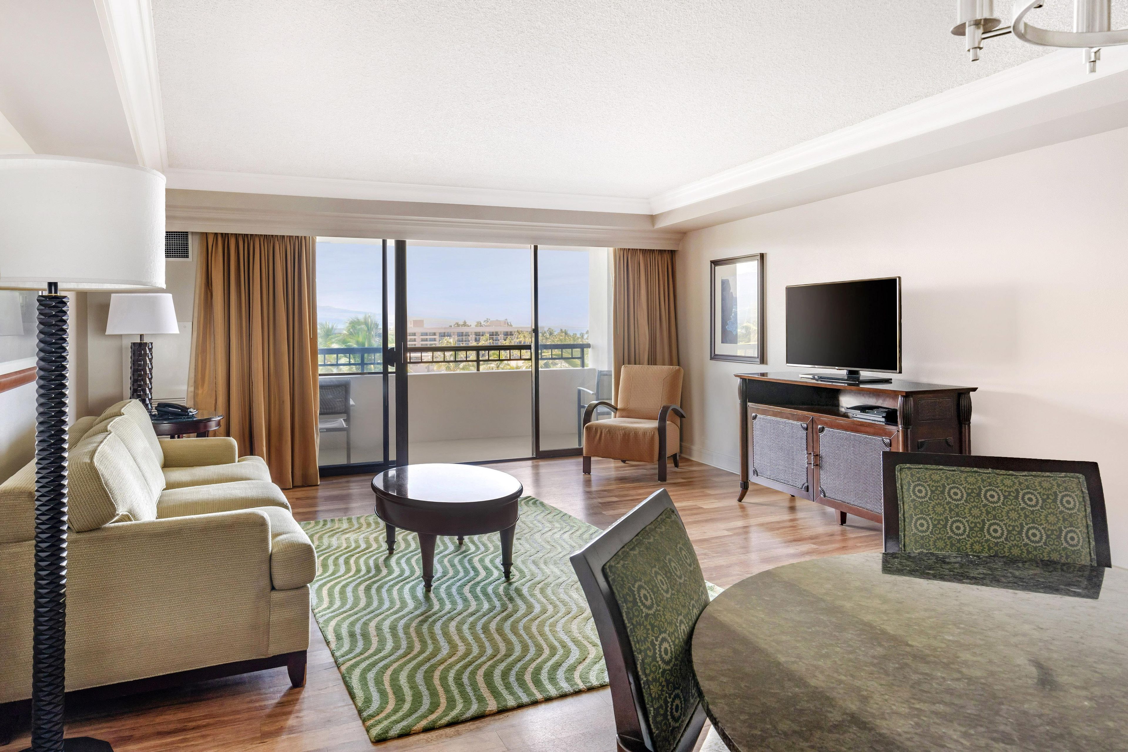 A hotel room with a sofa, armchair, TV, and a view of the city through large windows.