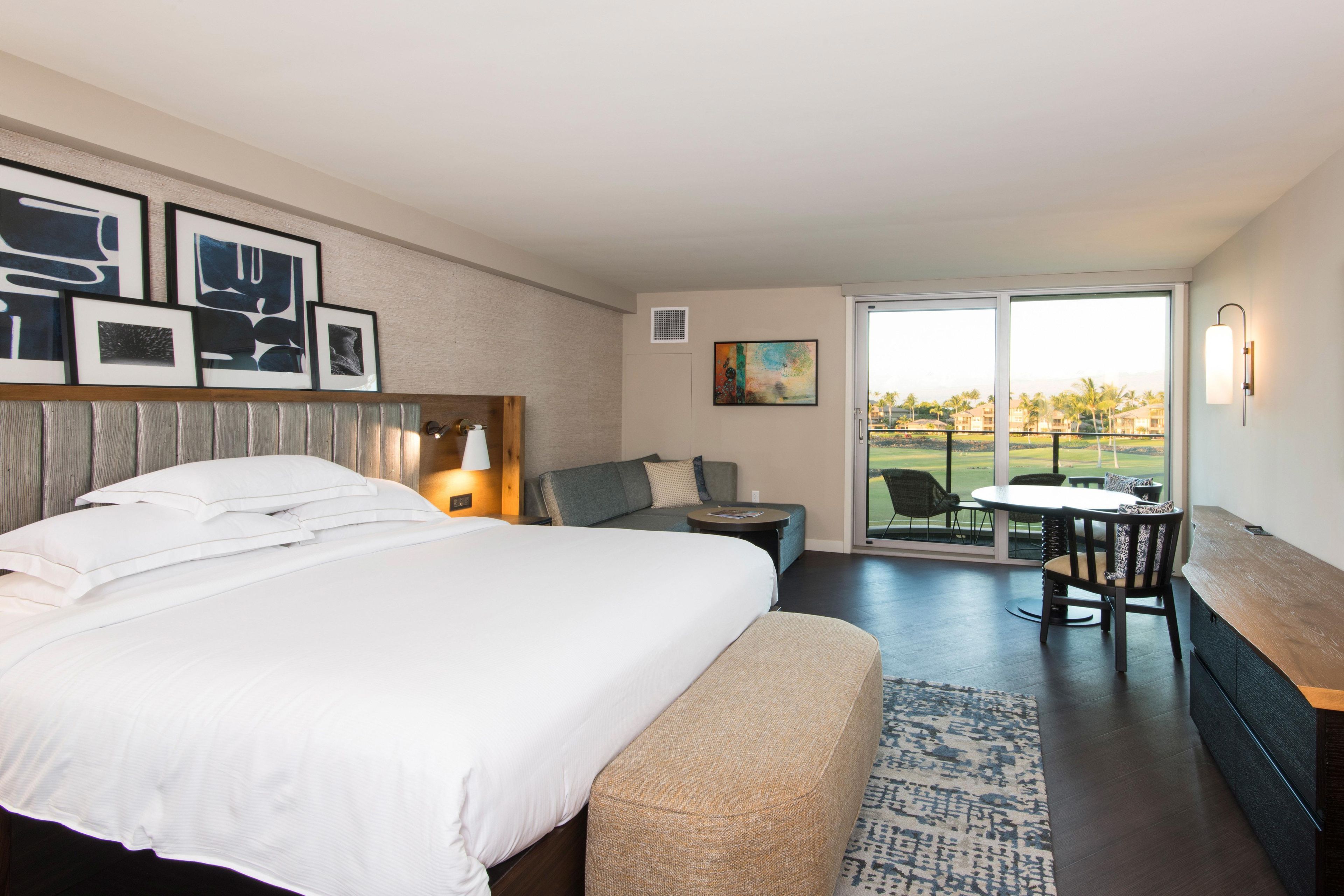 A hotel room with a large bed, a sofa, a small table, and a balcony with a view of the landscape.