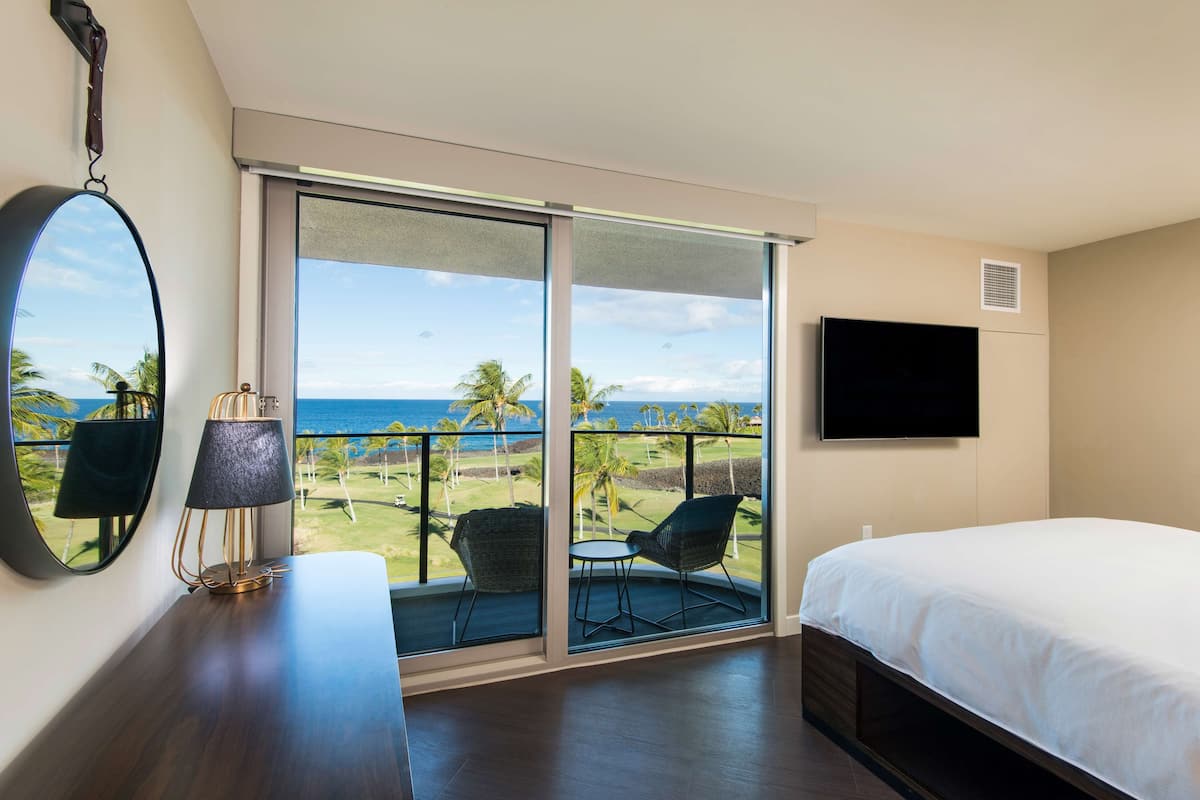 Une chambre d’hôtel dotée d’une grande porte-fenêtre coulissante donnant sur un balcon avec vue sur l’océan et les palmiers. La chambre comprend un lit, un téléviseur fixé au mur et une table avec des chaises.