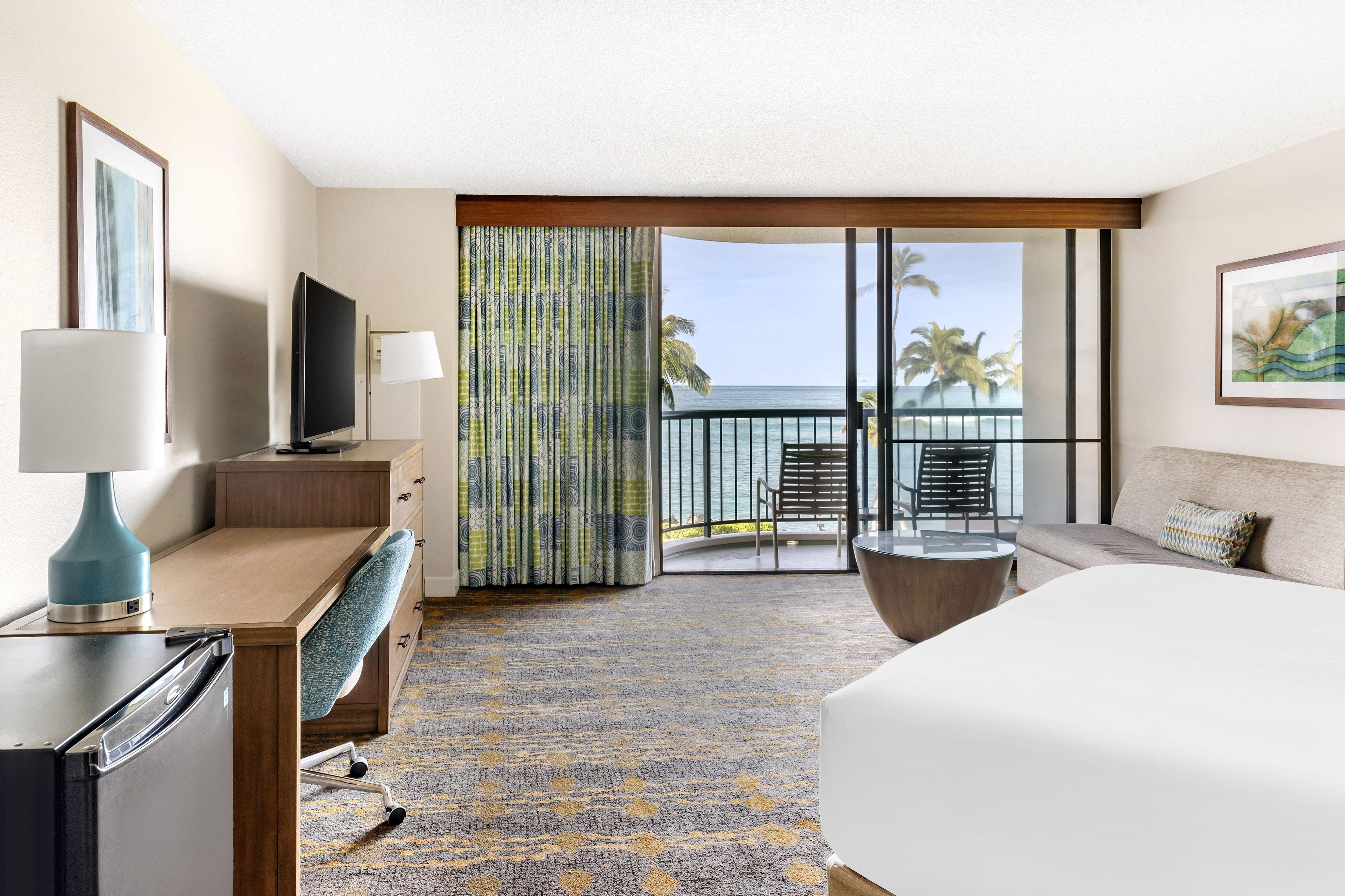 A hotel room with a bed, desk, TV, and a view of the ocean through a sliding door.