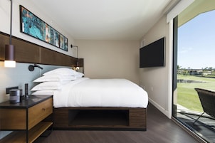 A modern hotel room with a large bed, a desk with a lamp, a flat-screen TV, and a view of a golf course.