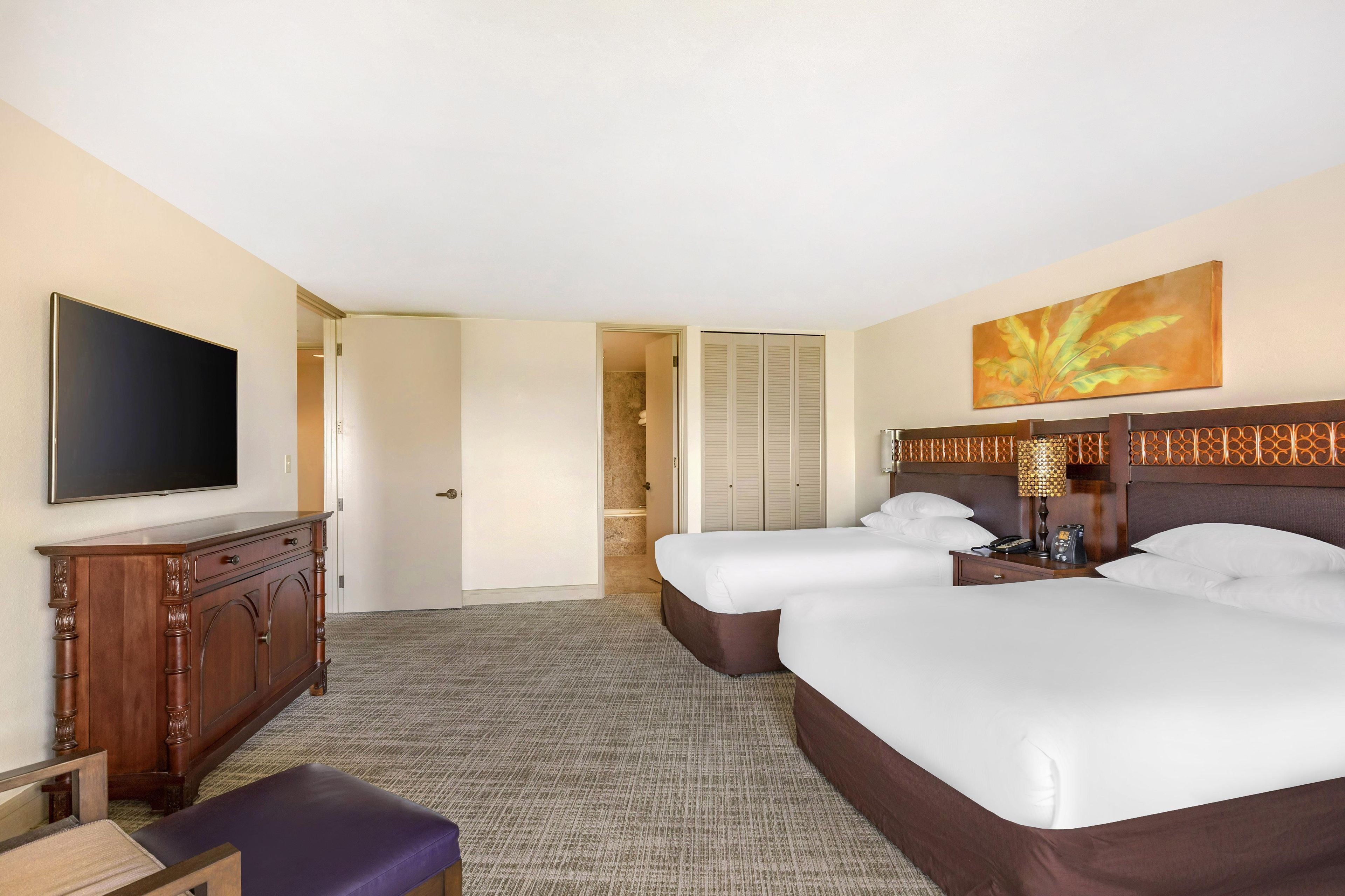 Quarto de hotel com duas camas, televisão, escrivaninha e banheiro visível por uma porta aberta.