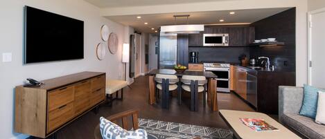 A modern living room with a flat-screen TV, a wooden cabinet, a dining area with a table and chairs, and a kitchen with dark cabinetry and appliances.
