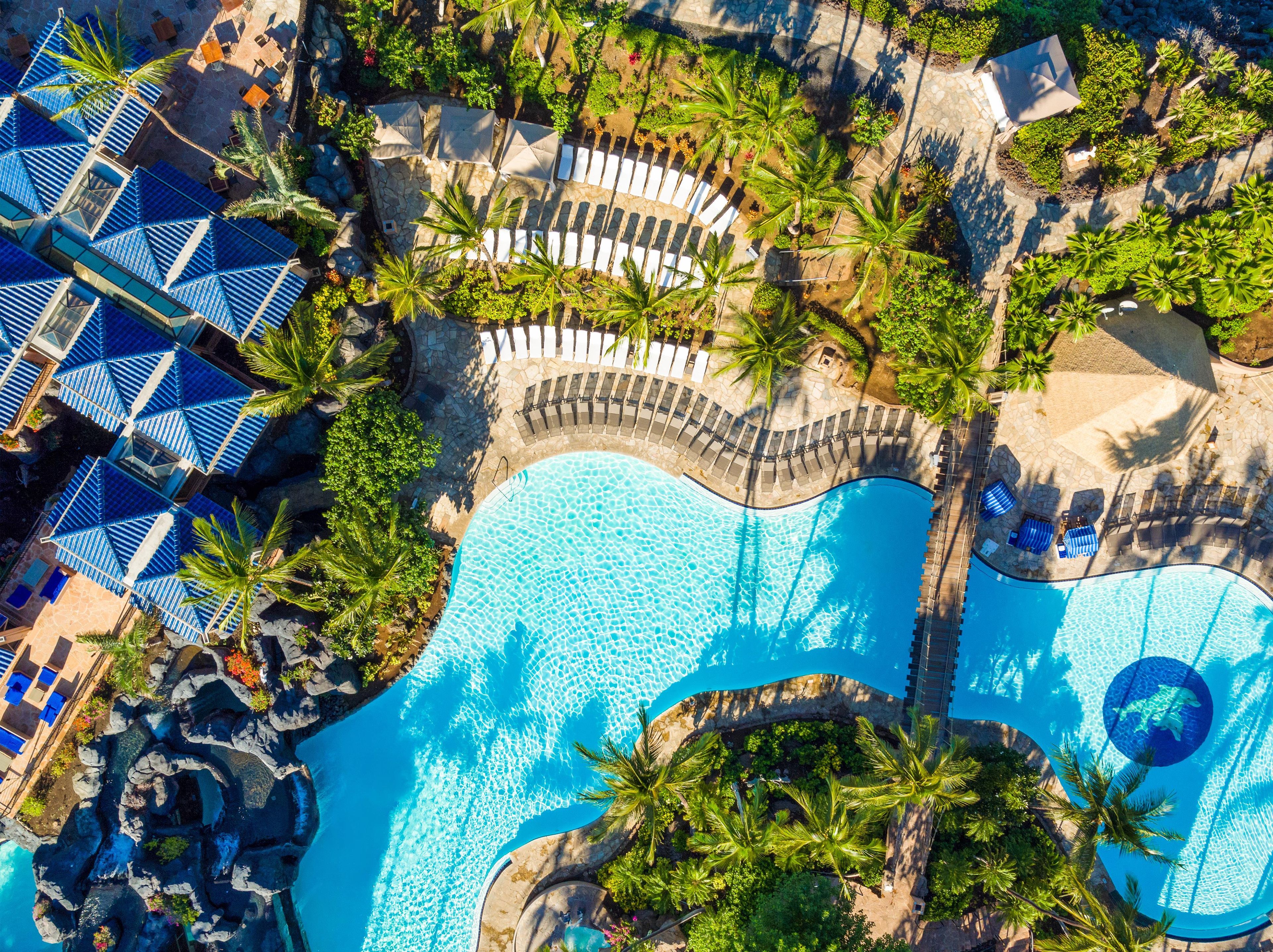 An aerial view of a resort with a large swimming pool, lounge areas, and palm trees.