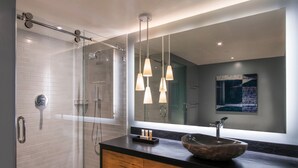 Een moderne badkamer met een glazen douchecabine, een houten wastafelmeubel met een grote waskom en hangende lampen.