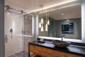 Une salle de bain moderne dotée d’une douche à parois de verre, d’un meuble-lavabo en bois avec un grand lavabo en forme de coupe et d’un éclairage suspendu.