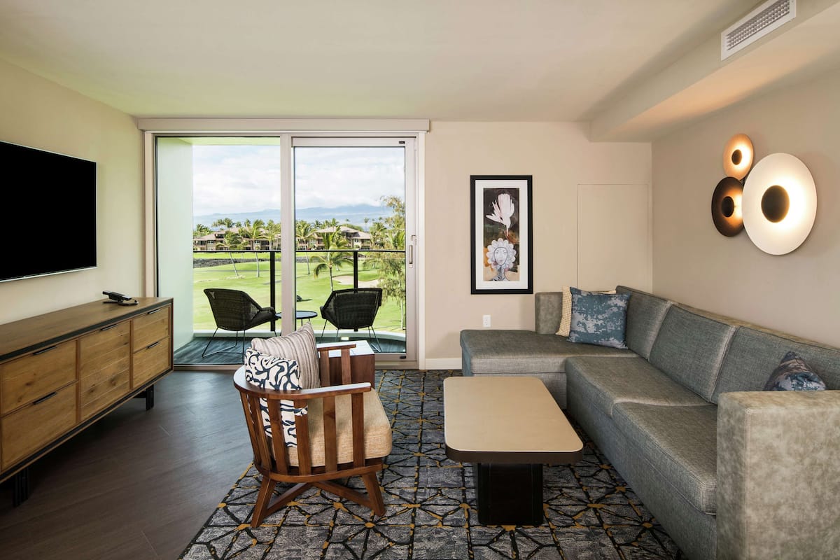 Un salon comprenant un canapé, un fauteuil et une table basse. Un téléviseur à écran plat est fixé au mur. Une porte-fenêtre coulissante donne accès à un balcon d’où l’on peut admirer un terrain de golf et des montagnes.