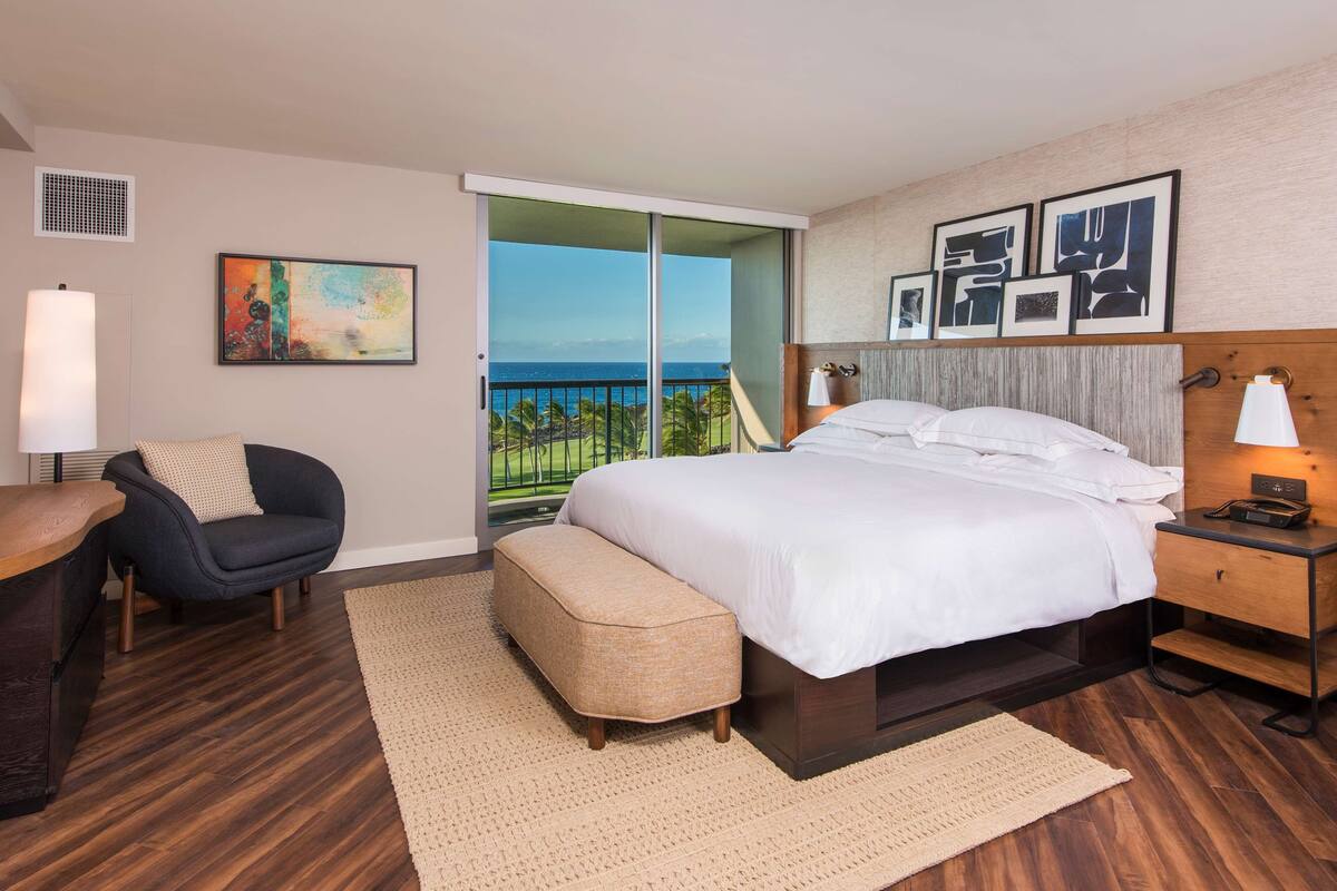 Une chambre d’hôtel moderne, dotée d’un grand lit, d’une chaise, d’un bureau et offrant une vue sur l’océan grâce à une porte coulissante.