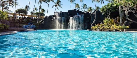 A swimming pool with a waterfall feature, surrounded by palm trees and a building with a tiled roof.