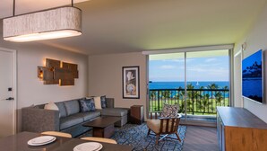 A modern living room with a sofa, dining table, and chairs, a large window with a view of the ocean, and a mounted artwork on the wall. - Hilton Grand Vacations Club Ocean Tower Waikoloa Village (Waikoloa)