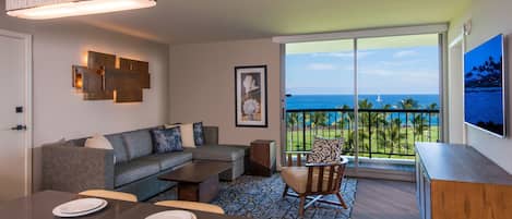 A modern living room with a sofa, dining table, and chairs, a large window with a view of the ocean, and a mounted artwork on the wall.