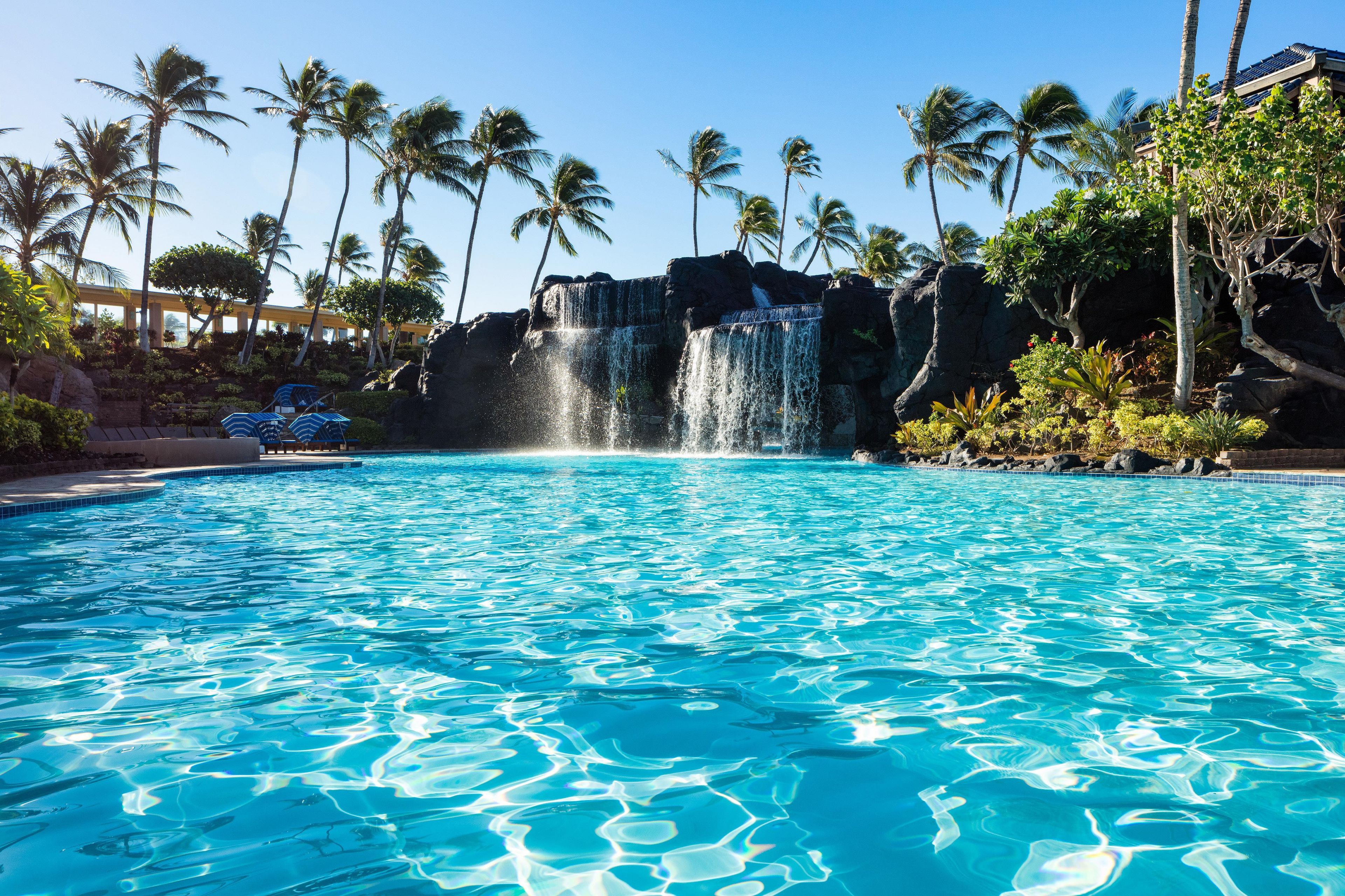 A swimming pool with a waterfall feature, surrounded by palm trees and a building with a tiled roof.