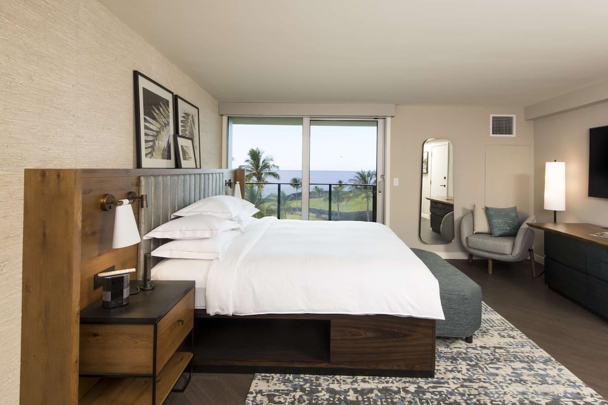 Une chambre d’hôtel moderne avec un grand lit, une table de chevet en bois, un bureau, une chaise, une télévision et un balcon offrant une vue sur l’océan.