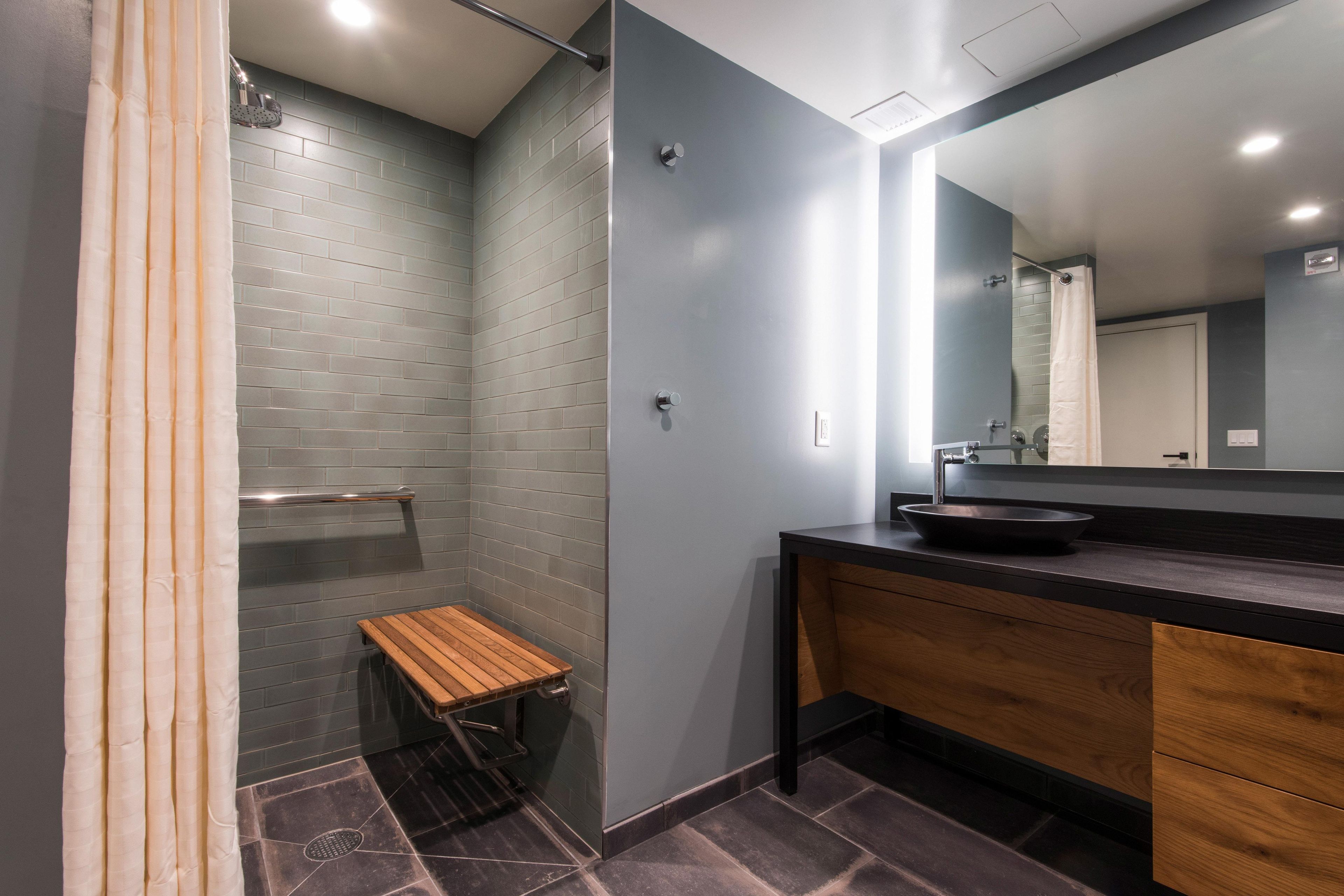 A modern bathroom with a shower, a wooden bench, a sink with a dark countertop, and a mirror.