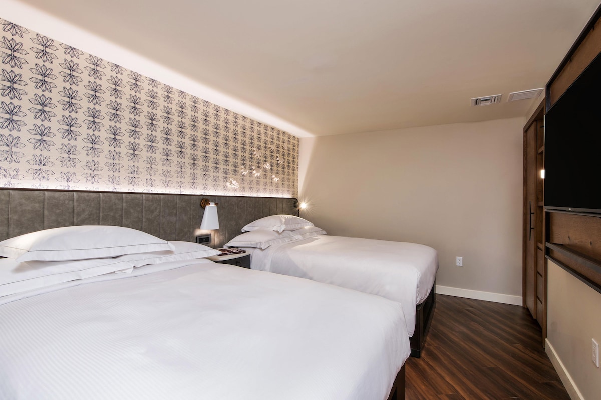 Une chambre d’hôtel avec deux lits, une tête de lit ornée d’un motif floral, une table de chevet avec une lampe et une armoire.