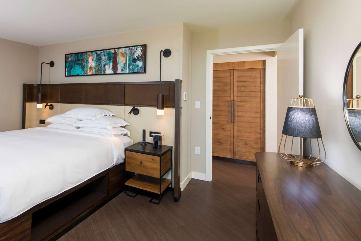 Une chambre d’hôtel comprenant un lit, une table de chevet, une lampe, une œuvre d’art et une armoire en bois.