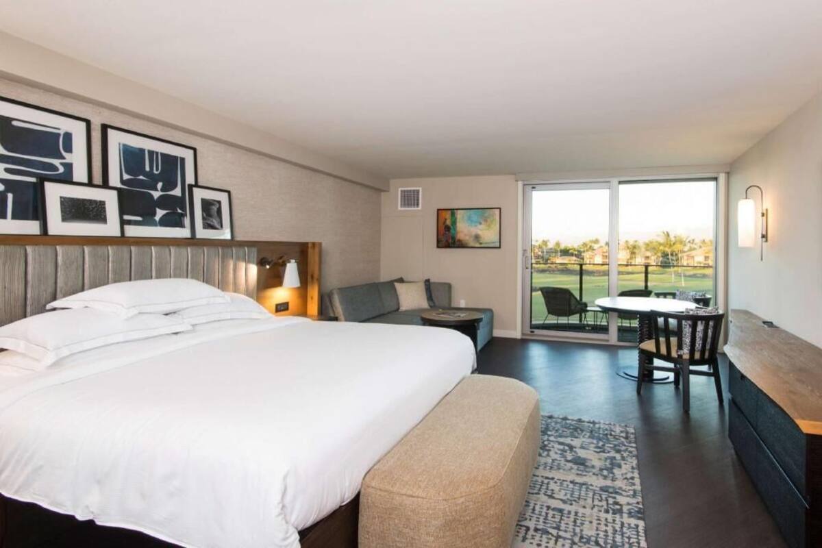 Une chambre d’hôtel dotée d’un grand lit, d’un coin salon avec une table et des chaises, d’un balcon offrant une vue agréable et d’œuvres d’art accrochées aux murs.