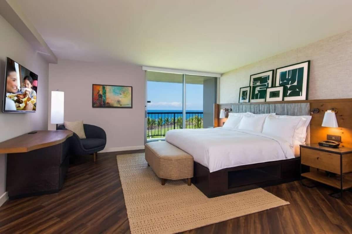 Une chambre d’hôtel équipée d’un lit, d’un bureau, d’une chaise, d’une petite table, d’une table de chevet et d’un balcon avec vue.