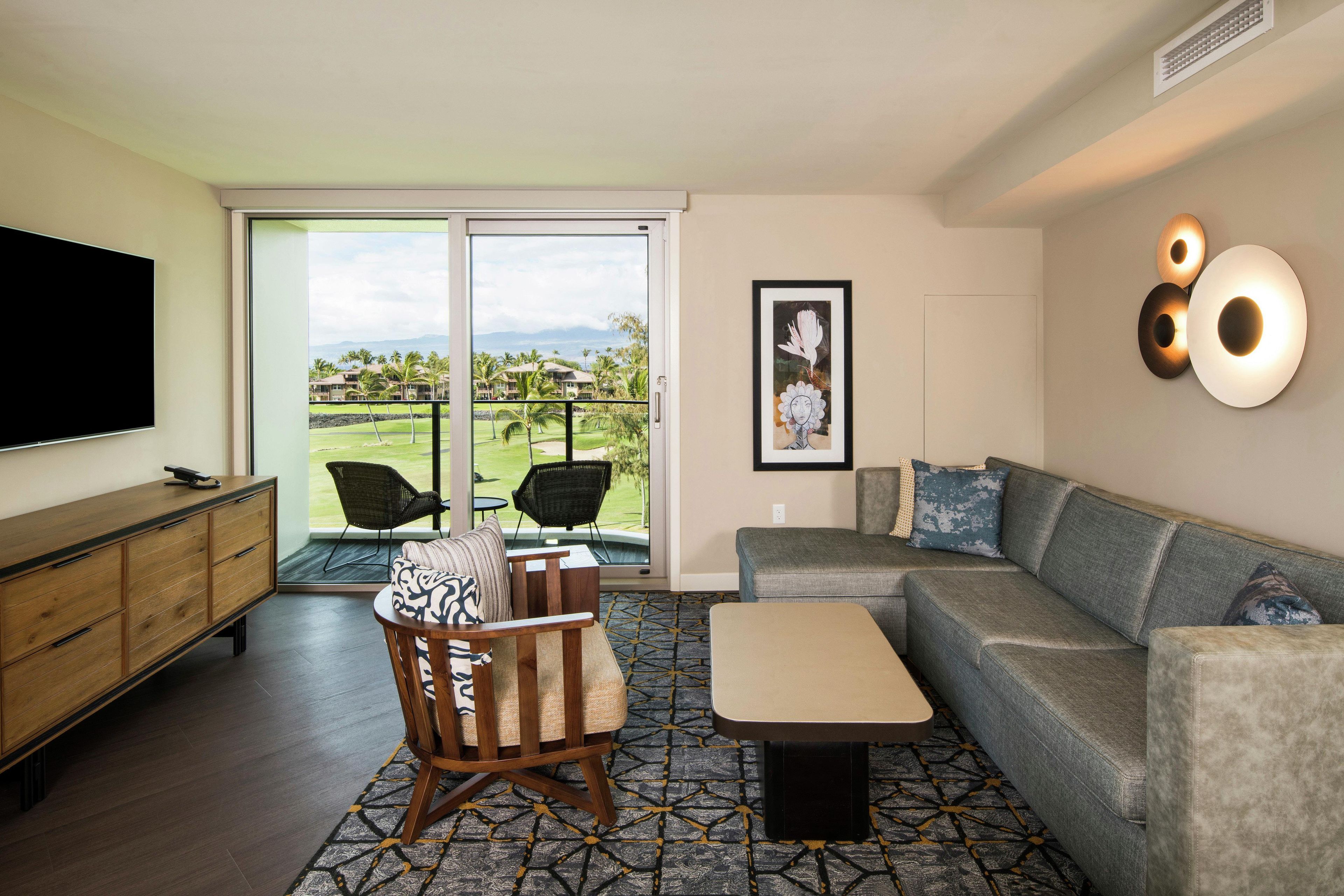 Un salon moderne avec un téléviseur à écran plat, un canapé, une table basse et un fauteuil. Une grande fenêtre offre une vue sur un terrain de golf et des immeubles.