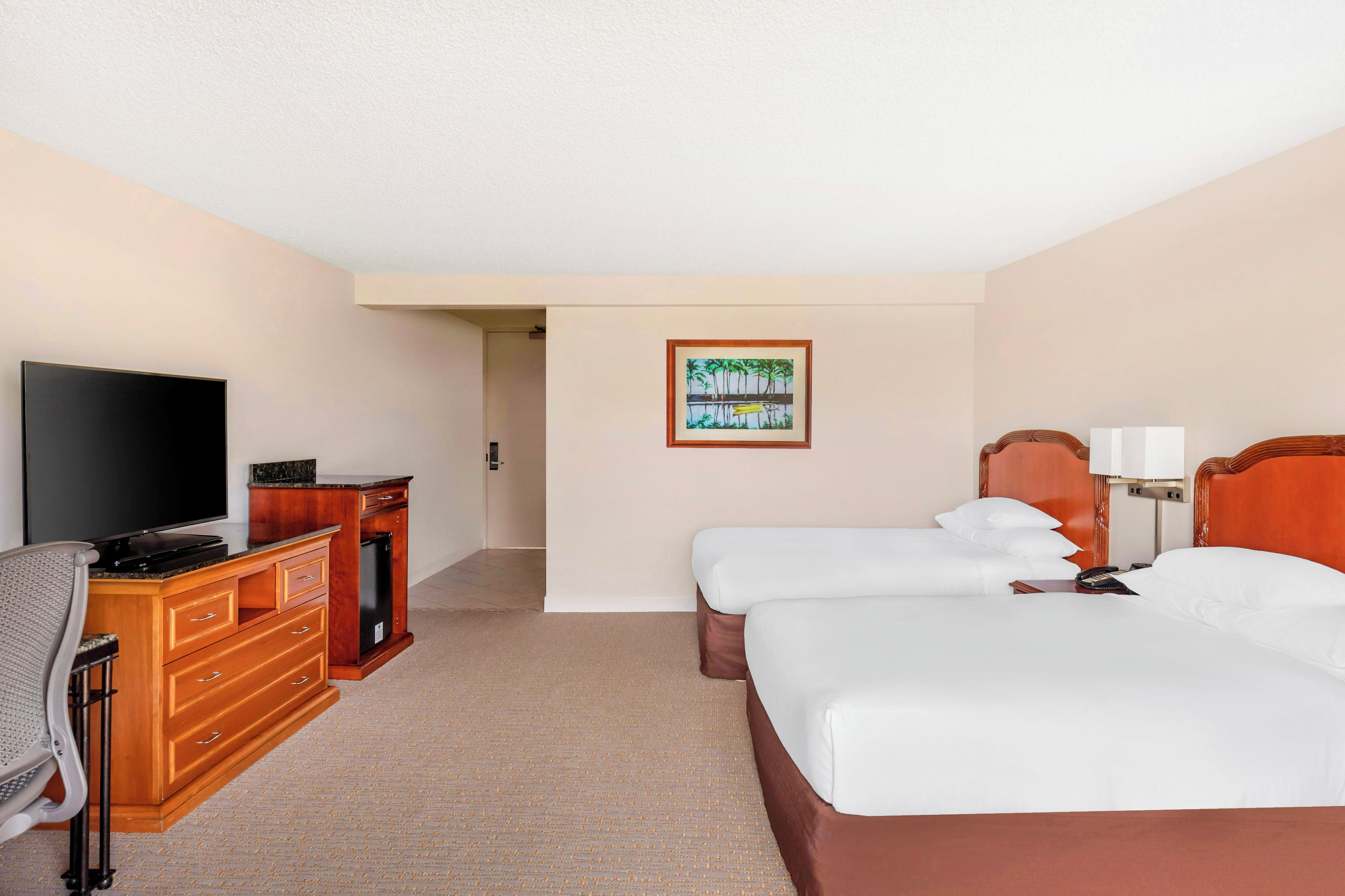 A hotel room with a flat-screen TV, a wooden dresser, two beds, a painting on the wall, and a lamp.