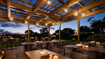 Een terras buiten met tafels en stoelen, een pergola met verlichting en uitzicht op de lucht.