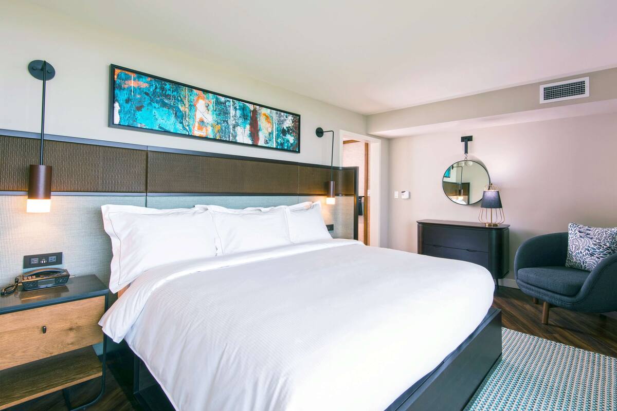 Une chambre d’hôtel moderne avec un grand lit, des tables de chevet, un bureau, une chaise, un miroir rond et une peinture abstraite aux couleurs vives accrochée au mur.
