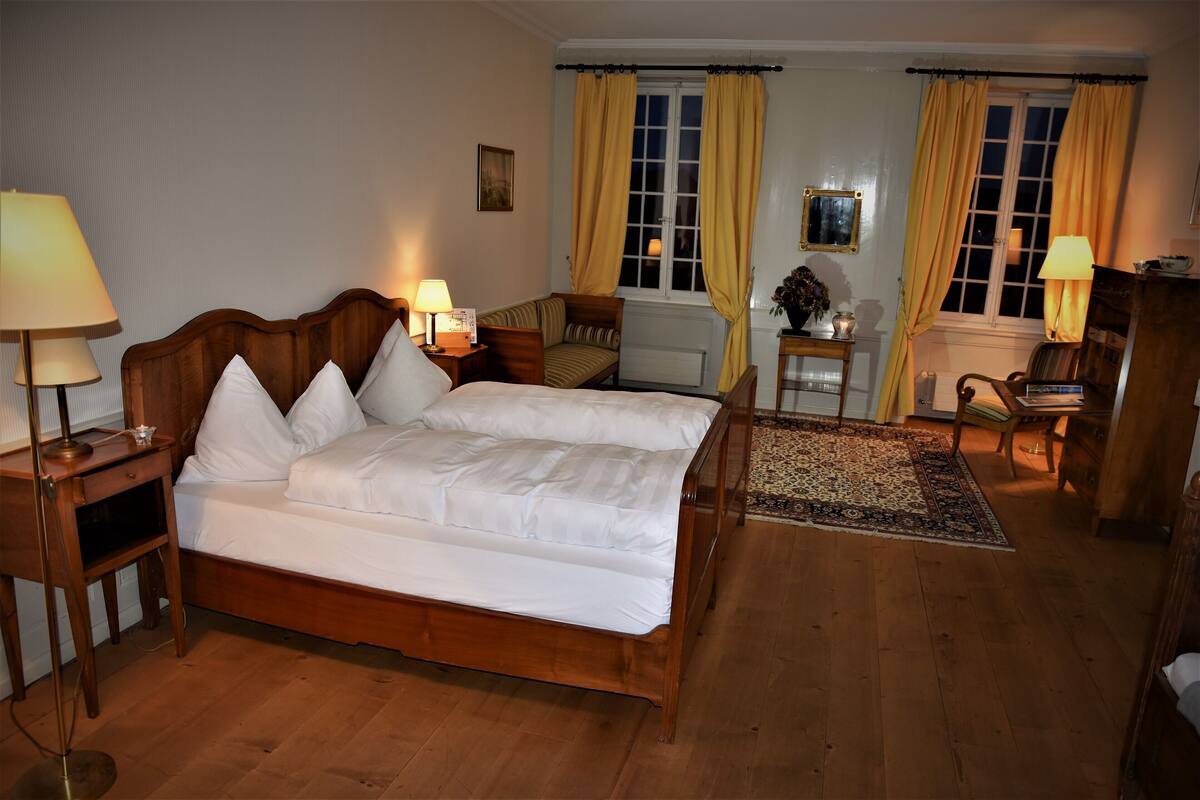 A bedroom with a wooden bed, bedside lamps, a small table with a lamp, a chair, a desk, and a window with curtains.