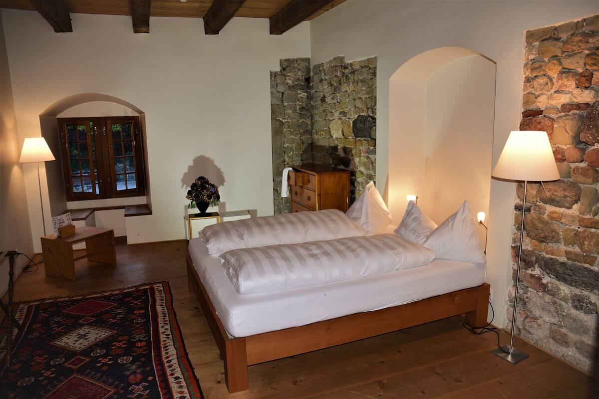 A bedroom with a wooden bed, a nightstand, a desk, a chair, a lamp, and a stone wall.