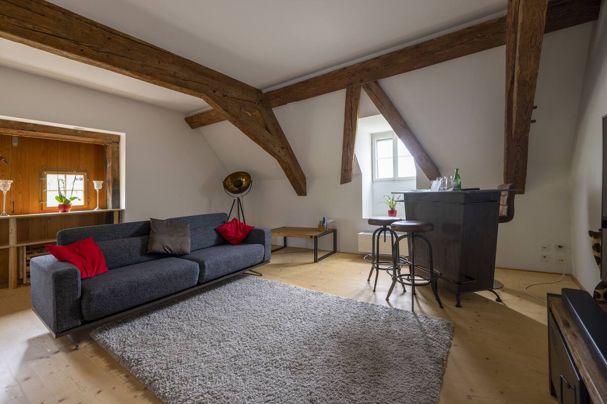 A loft-style room with exposed wooden beams, a gray sofa with red cushions, a small table, and a bar stool.