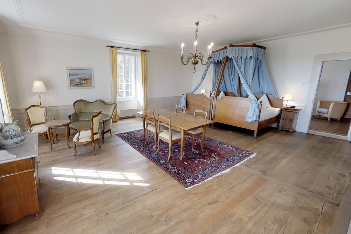 A bedroom with a four-poster bed, a dining table with chairs, a small table with a lamp, and a large mirror.