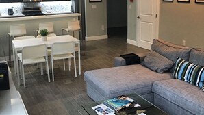 Una sala moderna con un sofá gris, una mesa de comedor con sillas, una cocina con armarios oscuros y un refrigerador de acero inoxidable.