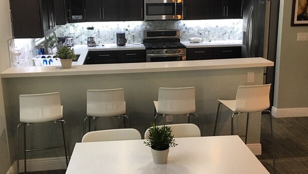 Una cocina moderna con armarios oscuros, un microondas de acero inoxidable y un horno empotrado. Hay una mesa de comedor blanca con cuatro sillas y una planta en maceta.