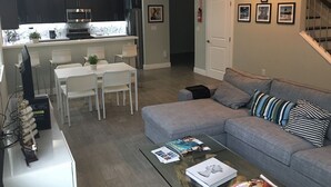 Una sala de estar moderna con un sofá gris, una mesa de centro de vidrio con revistas, un área de comedor con una mesa y sillas blancas, y una cocina con gabinetes oscuros y un horno empotrado.