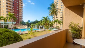 A balcony with a view of a swimming pool, palm trees, and a tall building in the background. - Ocho Rios Vacation - Apartment (Ocho Rios)