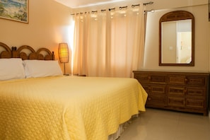 A bedroom with a large bed, wooden headboard, a bedside lamp, a mirror, and a wooden dresser. - Ocho Rios Vacation - Apartment (Ocho Rios)