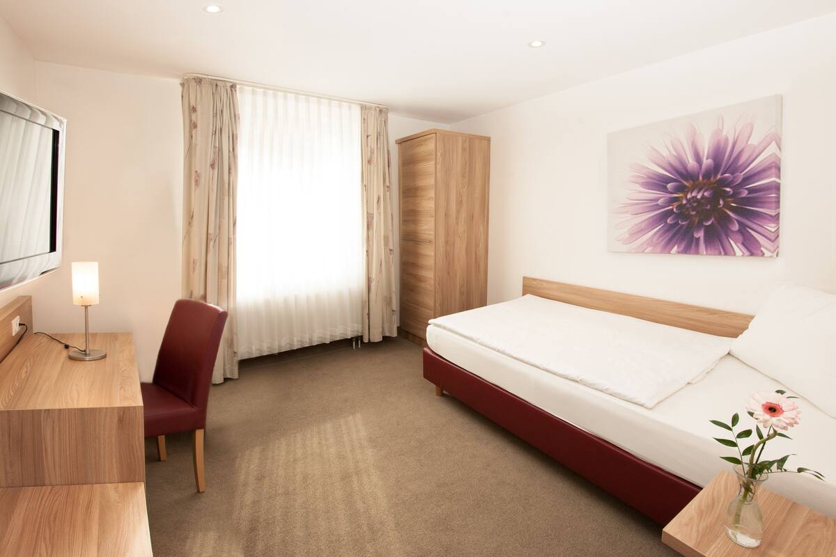 Une chambre d’hôtel avec un lit, un bureau, une chaise, une armoire et un tableau au mur.