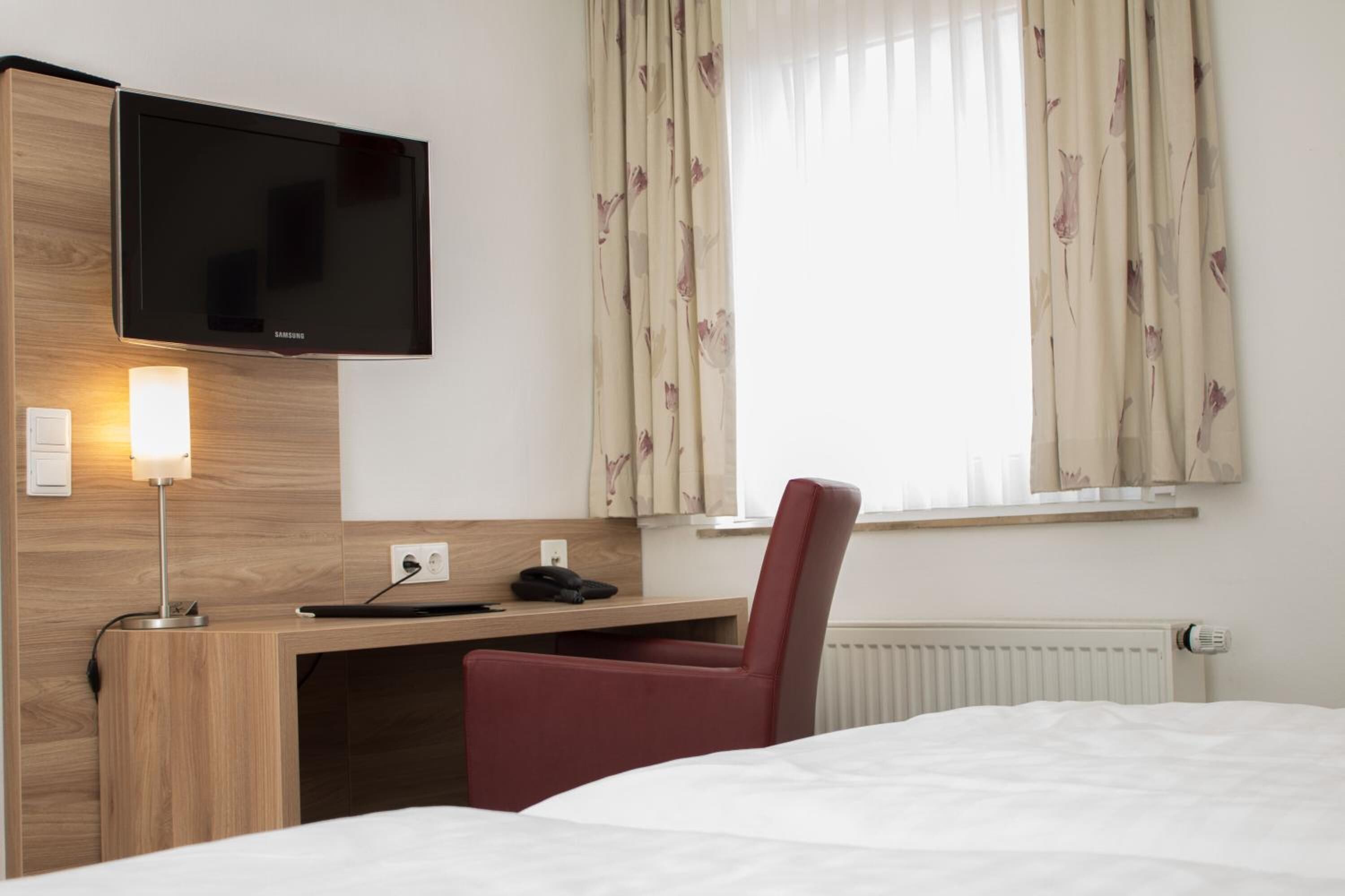 A hotel room with a bed, a desk with a chair, a flat-screen TV mounted on the wall, a lamp, and a window with curtains.