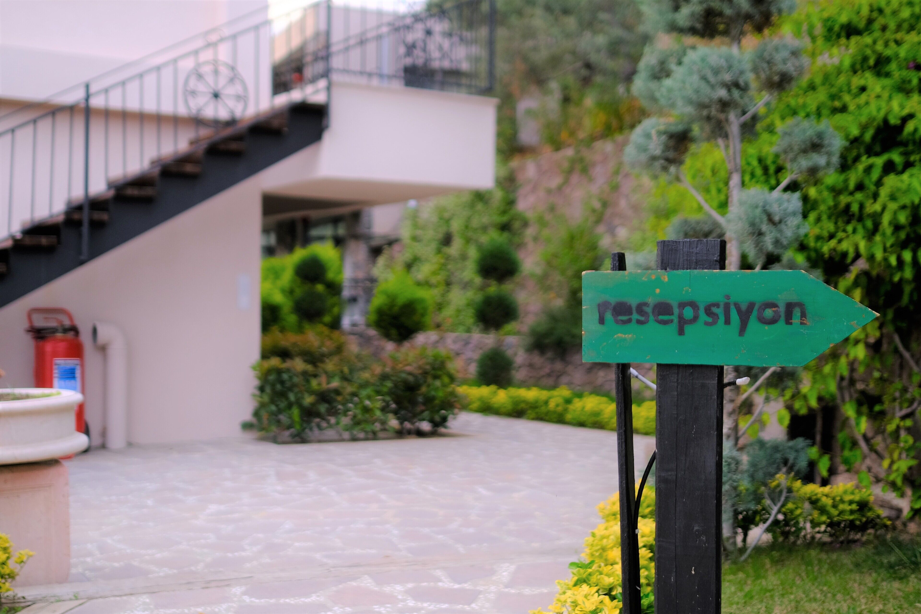 Ein Wegweiser mit dem Schriftzug "resepsiyon" weist auf einen Empfangsbereich, eine Treppe und einen angelegten Garten mit Feuerlöscher hin.