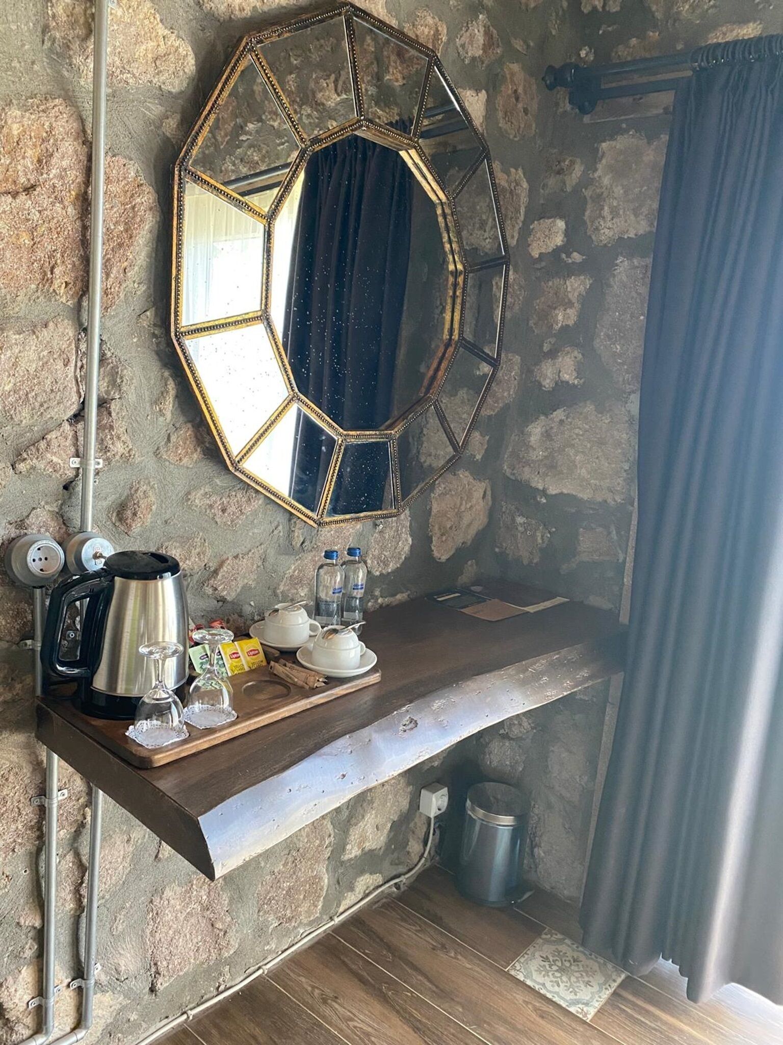 A rustic stone wall with a wooden shelf holding a kettle, glasses, and bottles. A hexagonal mirror hangs above the shelf.